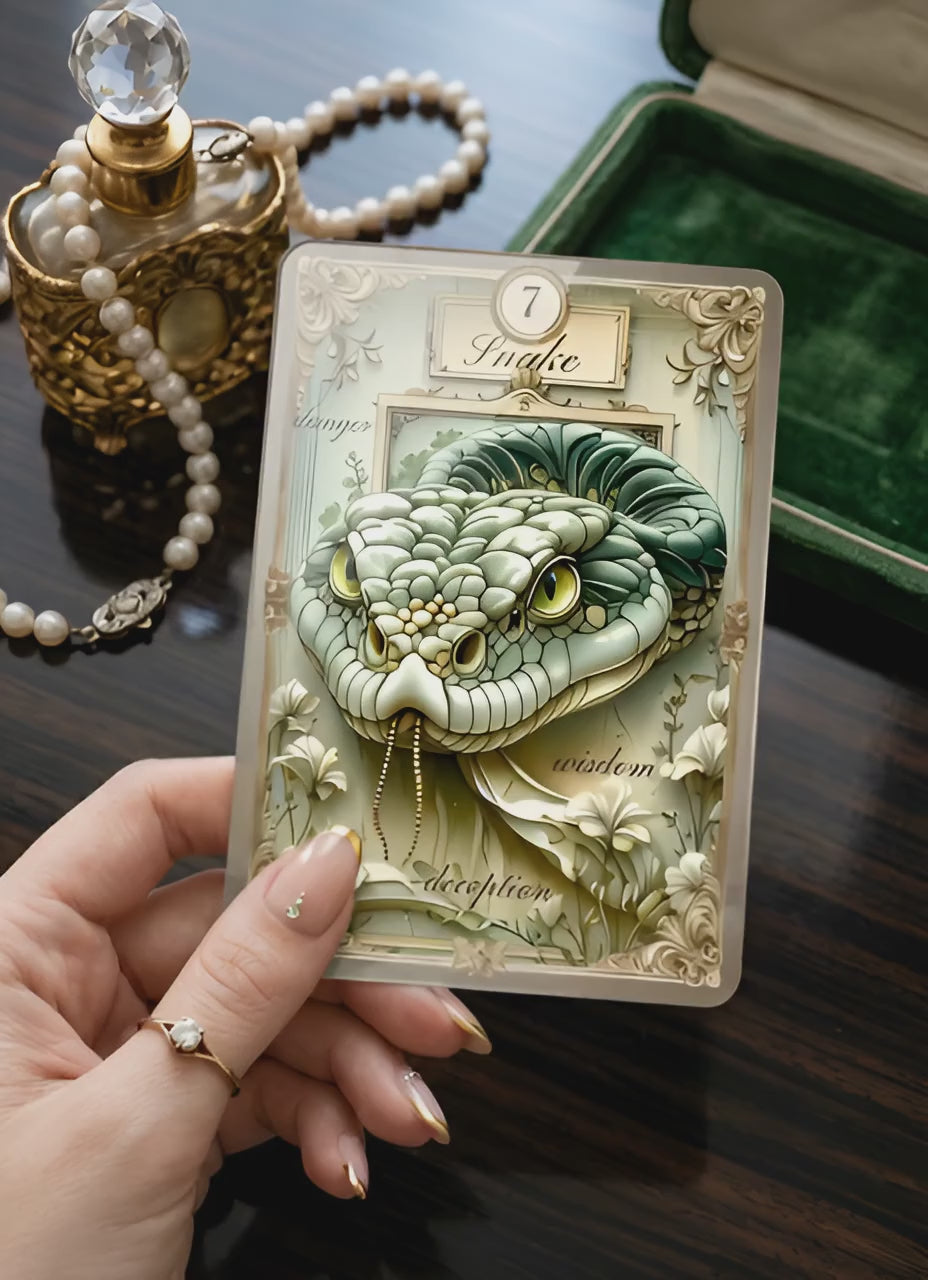 Hand holding Gilded Dreams Lenormand Snake card — glossy lamination macro shot with vintage perfume bottle, pearl necklace and green velvet jewelry box, handmade vintage oracle MardocCraft